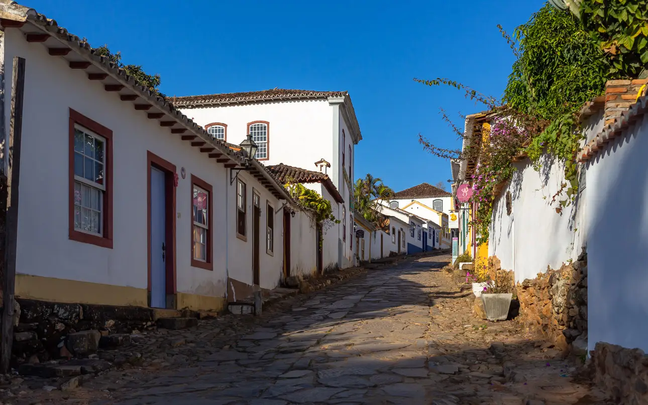 Weiße Häuser in einer engen Straße Eine enge Straße mit weißen Häusern und roten Fensterrahmen. Die Straße ist gepflastert und von Pflanzen umgeben.