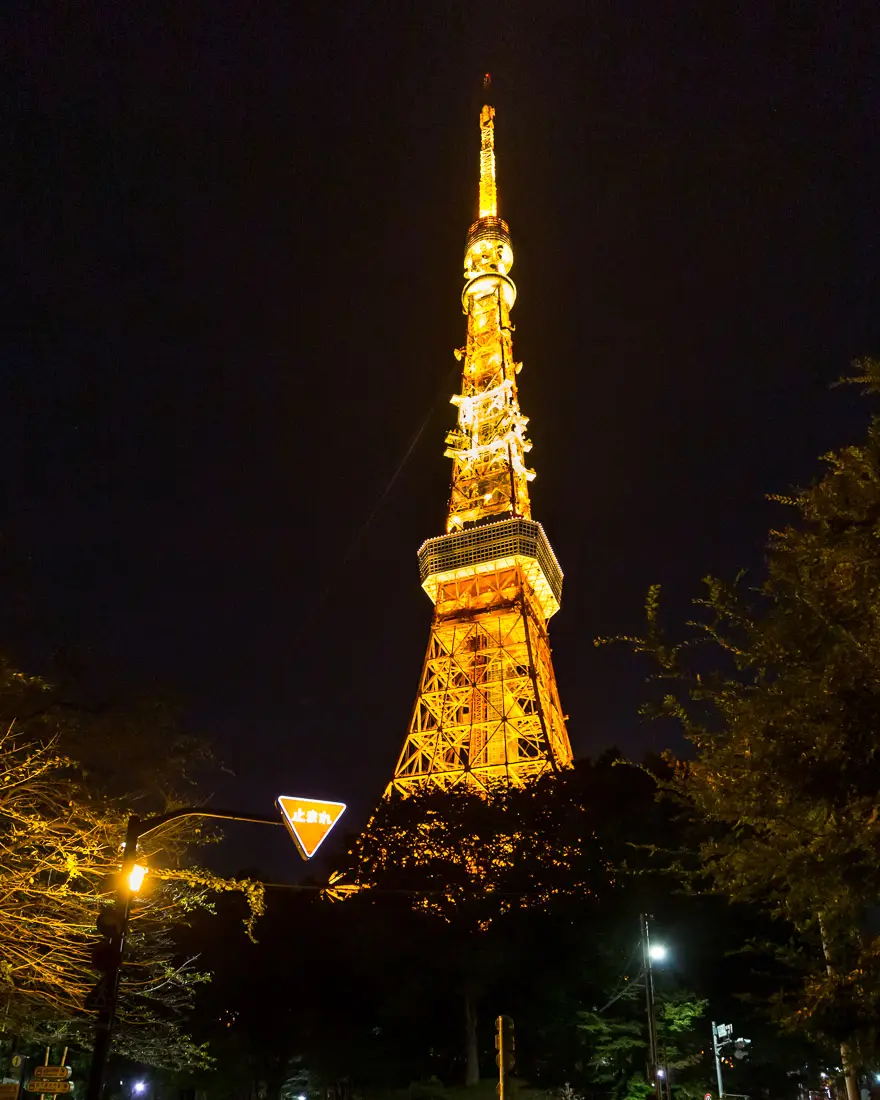 Tokio Turm bei Nacht Ein hoher, goldener Turm in der Nacht, umgeben von Bäumen und Straßenlaternen.