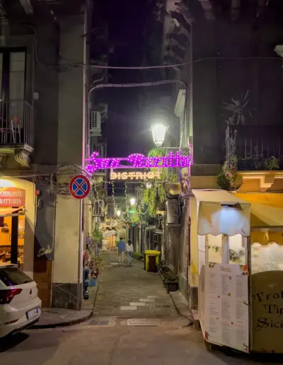 Eine enge Straße in Italien bei Nacht, beleuchtet von Straßenlaternen und bunten Lichtern.