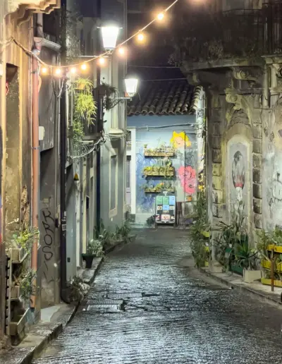 Eine enge Straße in einer Stadt bei Nacht, die von Lichterketten erleuchtet wird. Die Häuser sind alt und haben Graffiti und Pflanzen.