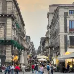 Menschen gehen durch eine Straße mit historischen Gebäuden und Cafés. Die Straße ist belebt und die Gebäude haben eine rustikale Atmosphäre.