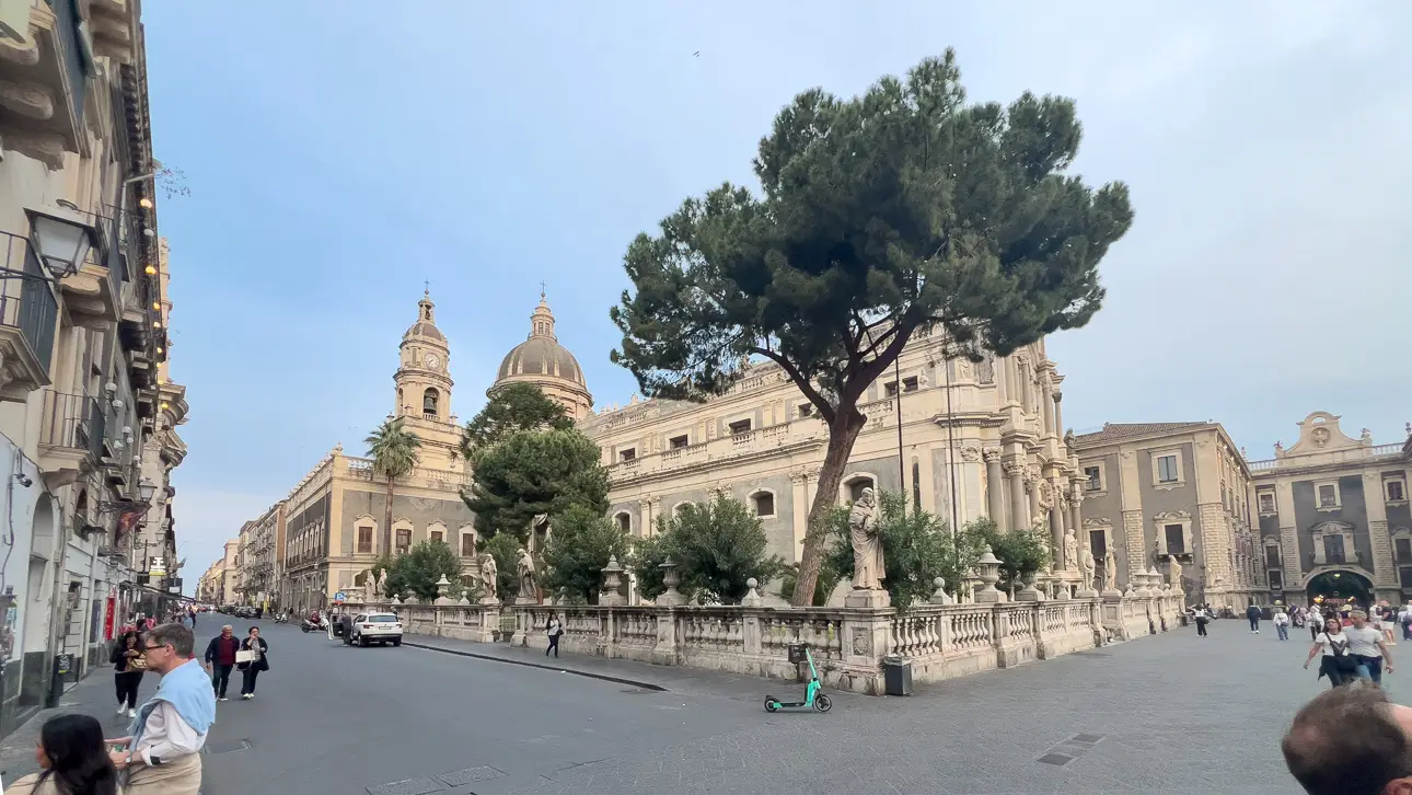 Ein großes historisches Gebäude mit zwei Türmen und einer Kuppel, umgeben von Bäumen und Menschen auf der Straße.