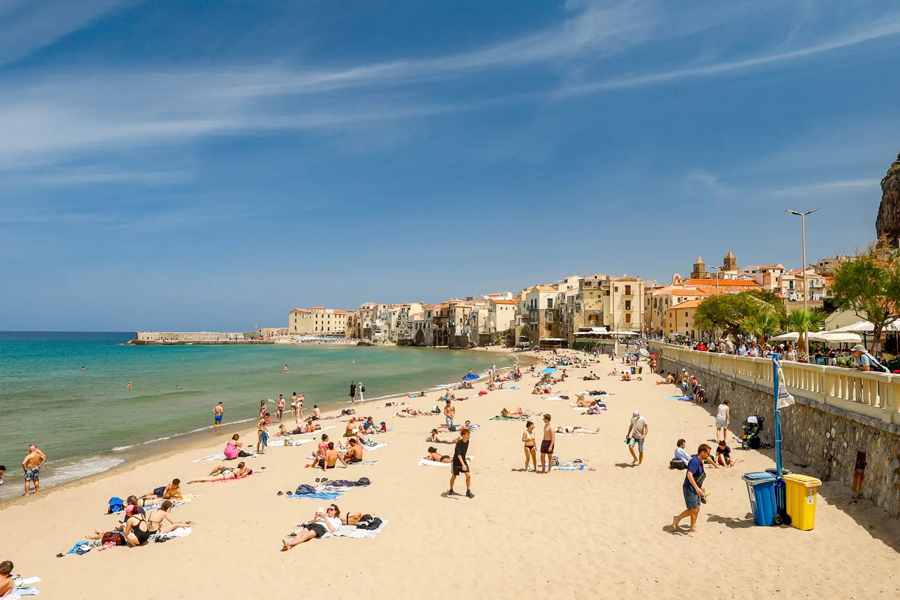 Ein Strand mit vielen Menschen, die sich sonnenbaden und im Wasser schwimmen. Im Hintergrund sind Gebäude und ein klarer Himmel.