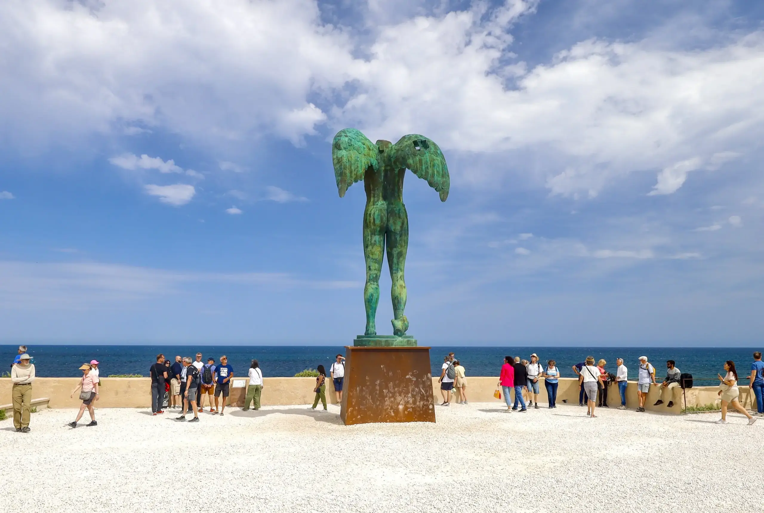 Ein großes, grünliches Standbild mit ausgebreiteten Armen vor einer großen Gruppe von Menschen.